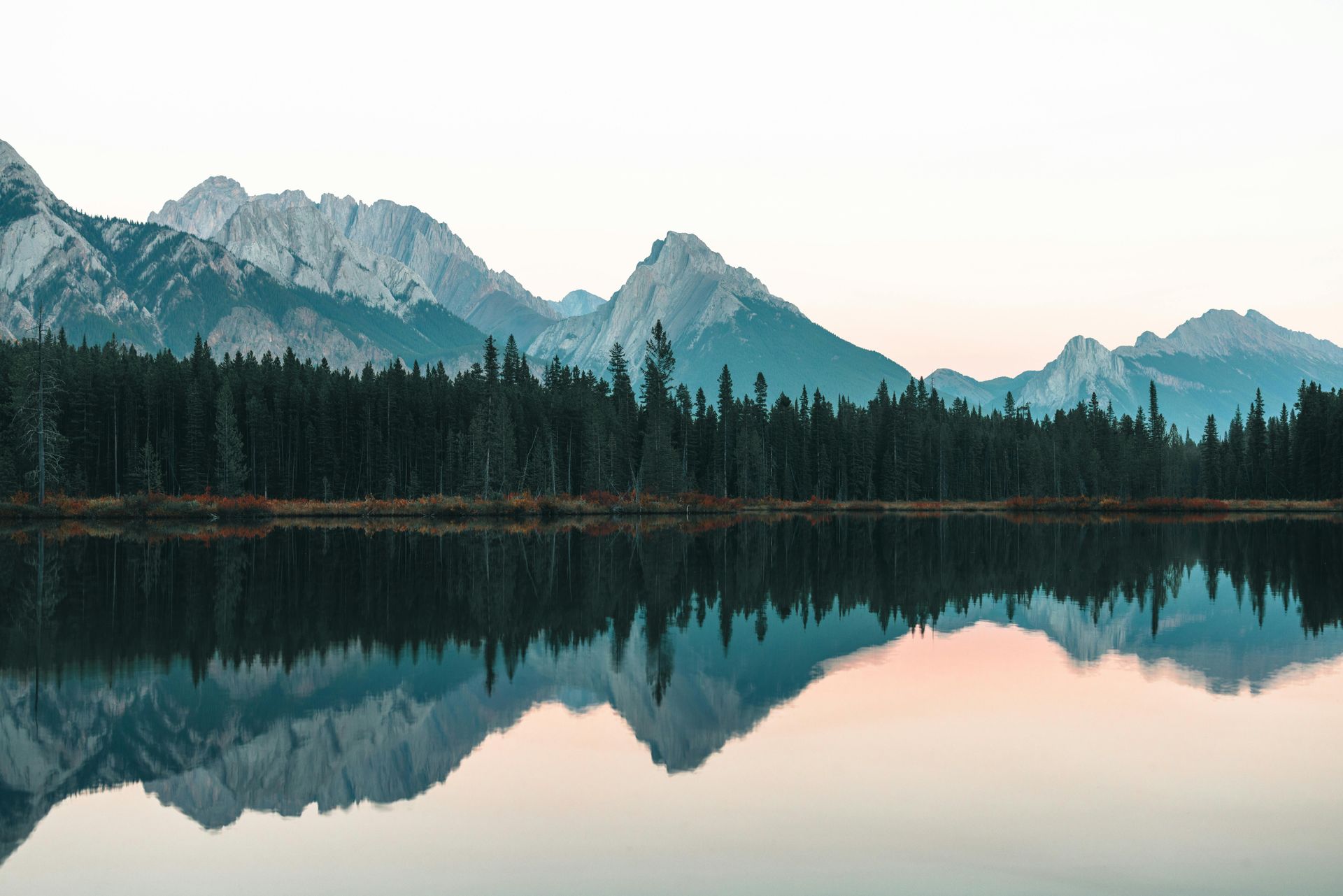 Mountains reflected in a calm lake, with a dark forest along the shoreline under a soft, pastel sky.
