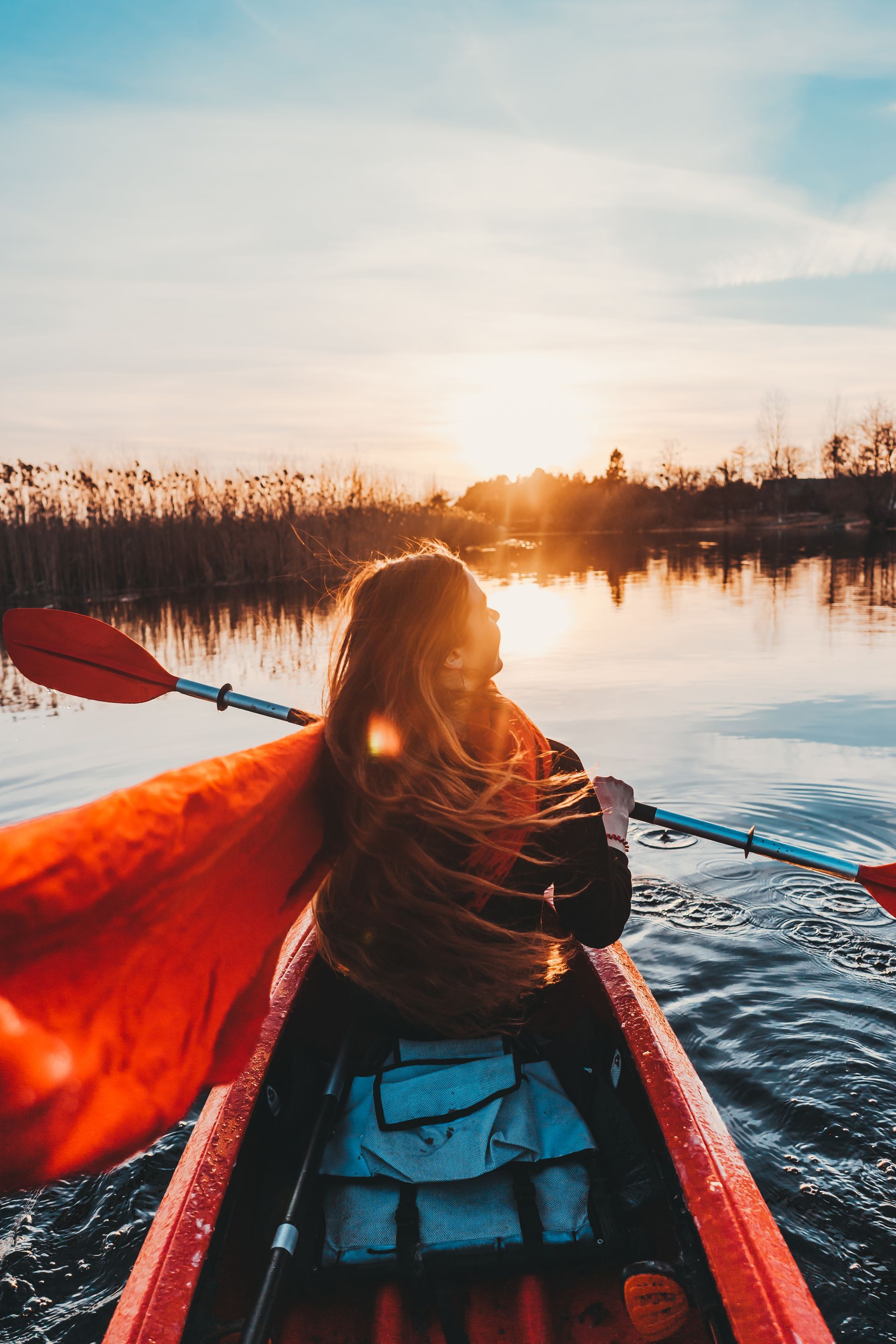 Woman kayaking on a lake at sunset, hair blowing, wearing a red scarf, reeds in the background.