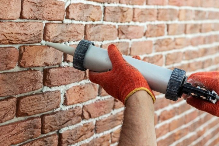 Gloved hands using a caulking gun to seal gaps in a brick wall