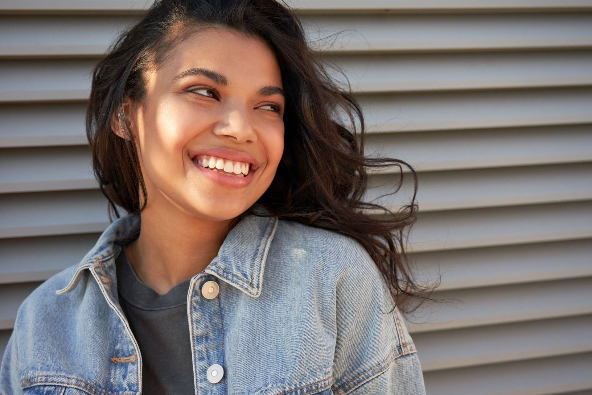 A woman in a denim jacket is smiling in front of a wall.