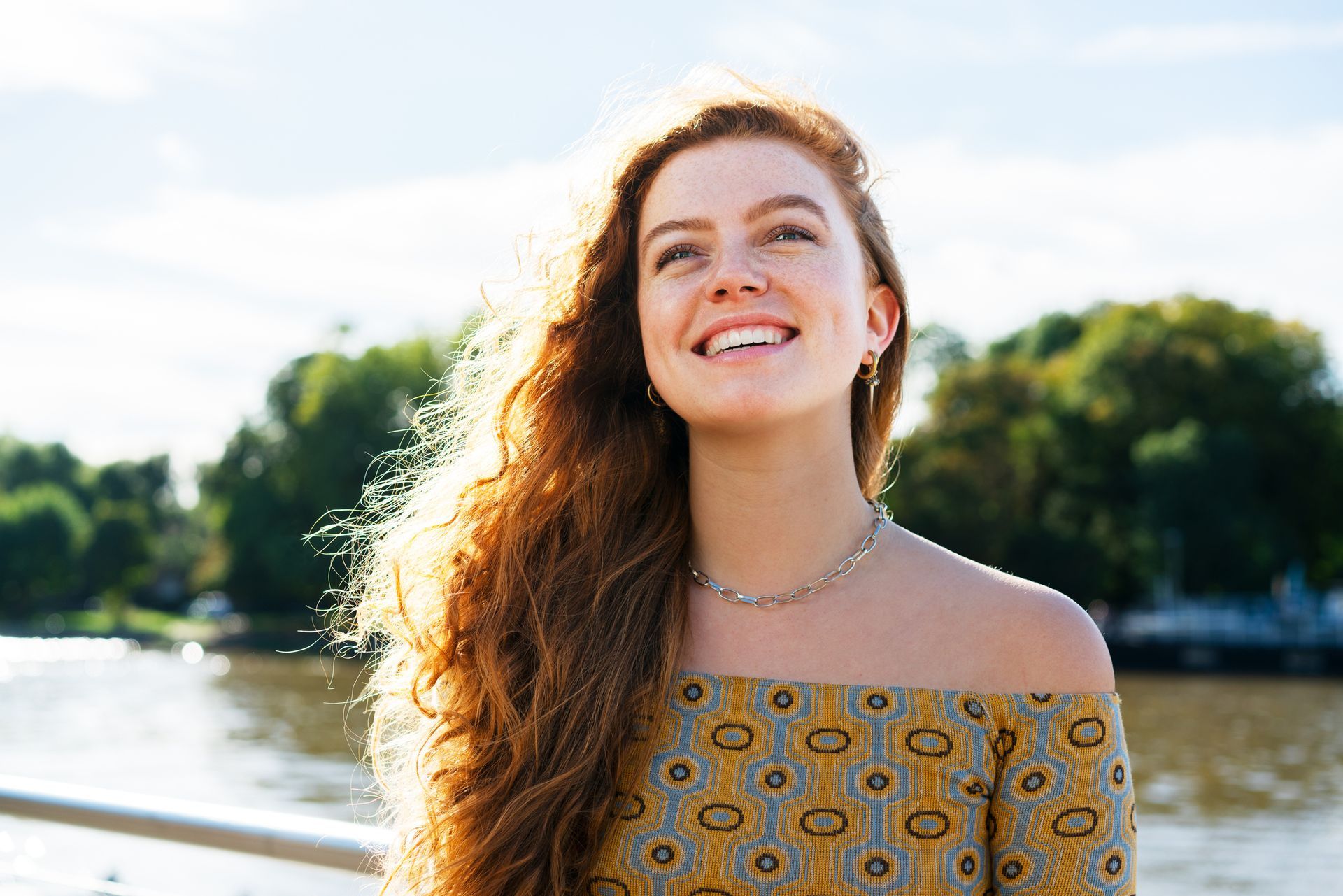 A woman with long red hair is smiling in front of a body of water.