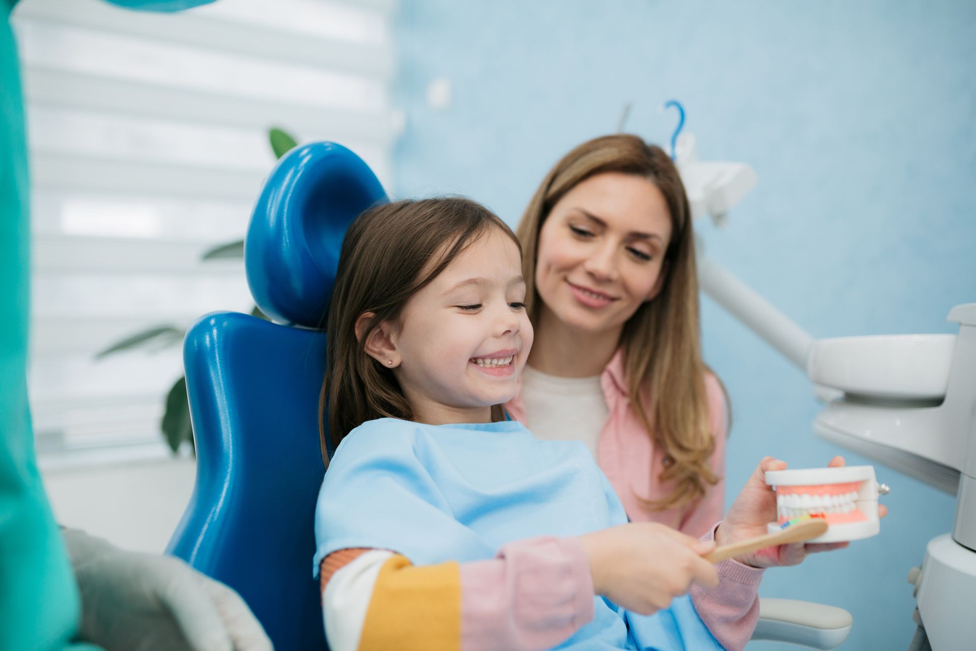 A woman is teaching a little girl how to brush her teeth in a dental office.
