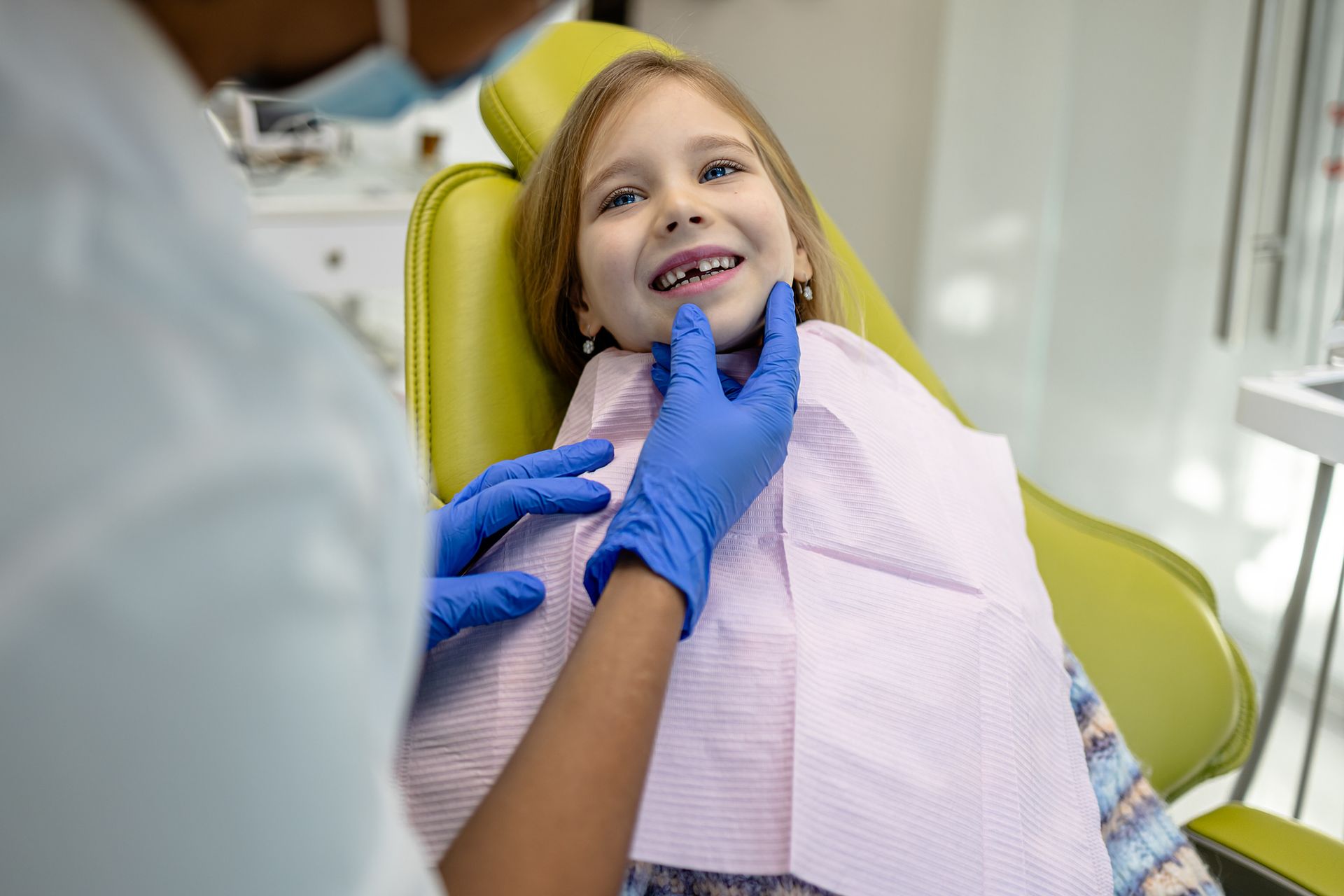 A dentist is sitting next to a little girl in a dental chair looking at a mirror.