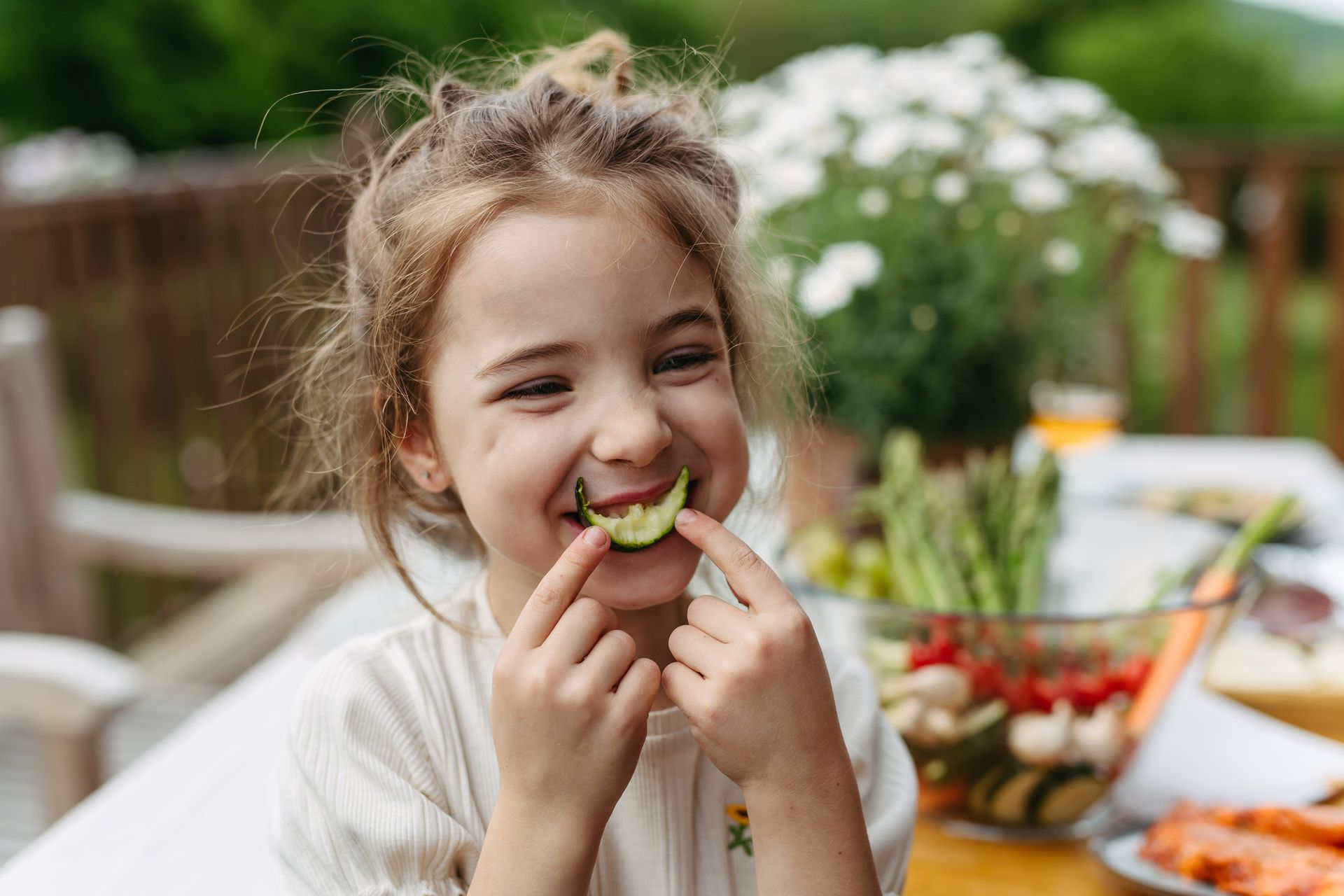 A little girl is eating a cucumber at a table.