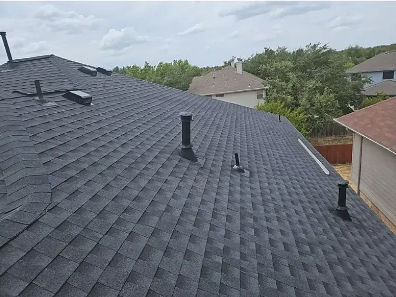 Dark gray asphalt shingle roof on a house, various vents, cloudy sky, green trees.