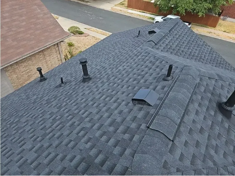 Dark gray shingle roof with black vents and a portion of a house visible in the background.