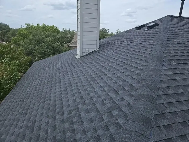 Dark gray shingle roof with a white chimney against a cloudy sky and green trees.