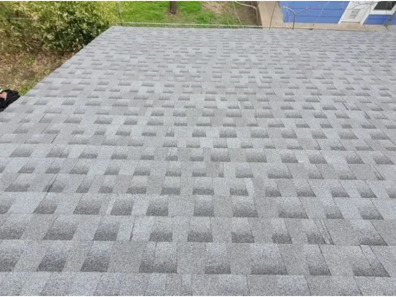 Gray asphalt shingle roof, angled view. Shingles in a pattern. Green grass and blue building visible.