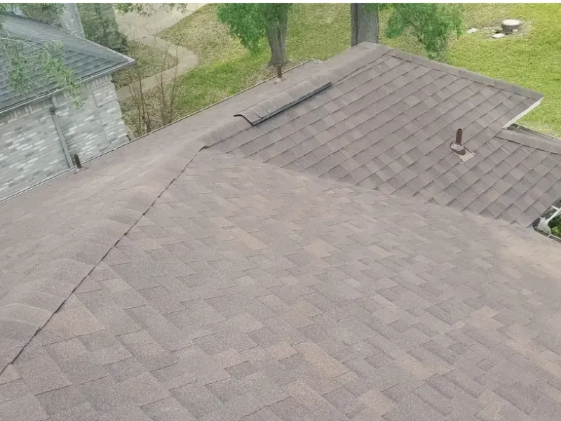 Brown asphalt shingle roof on a house, angled view. A vent and an adjacent building are visible.