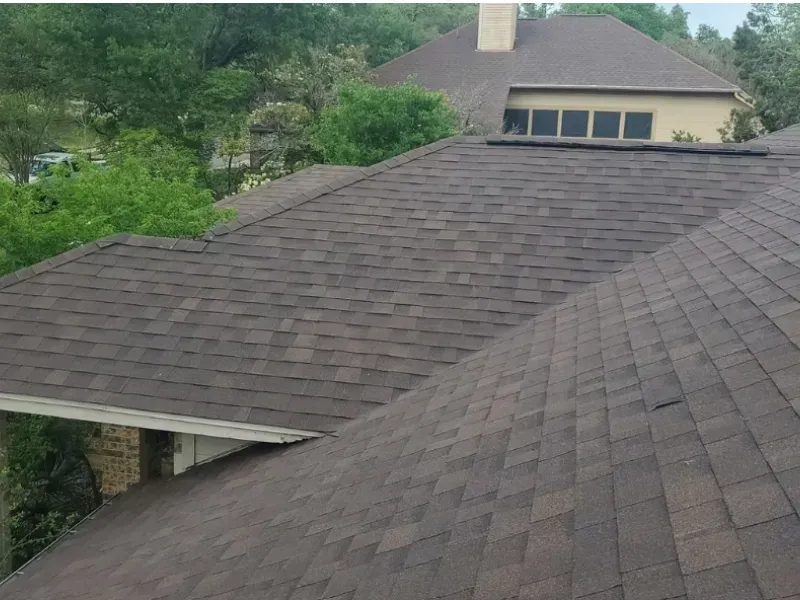 Brown asphalt shingle roof on a house, angled view. Surrounded by green trees and visible windows.