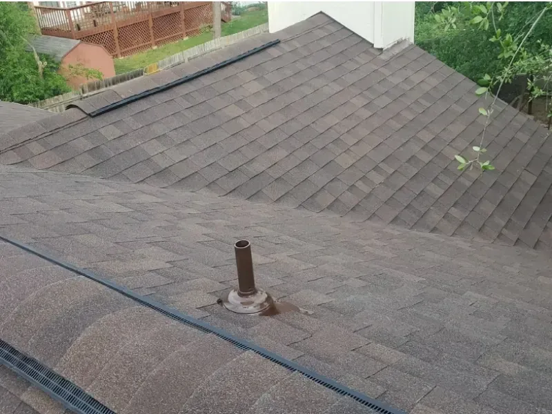 Dark gray shingled roof with a vent pipe and black flashing. Background shows a wooden fence and greenery.