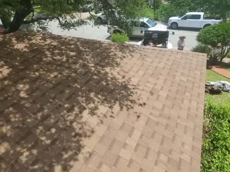 Brown shingle roof with tree shadows; workers on the roof near parked white vehicles on a sunny day.