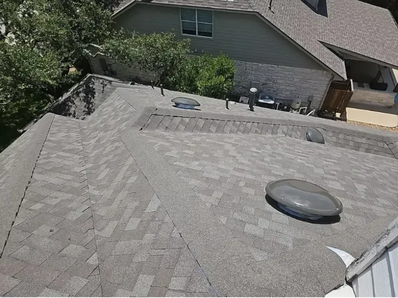 Overhead view of a gray asphalt shingle roof with vents, set against a background of a house and trees.