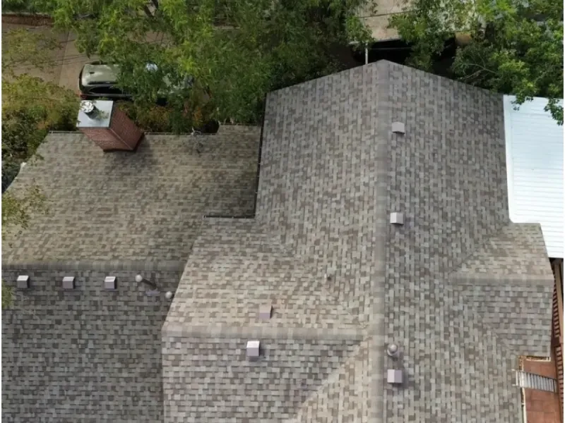 Overhead view of a gray asphalt shingle roof with vents and a chimney against a background of trees.