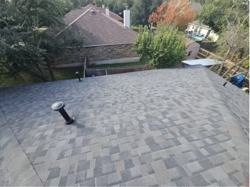 Overhead view of a gray asphalt shingle roof with a vent pipe and nearby houses with trees.