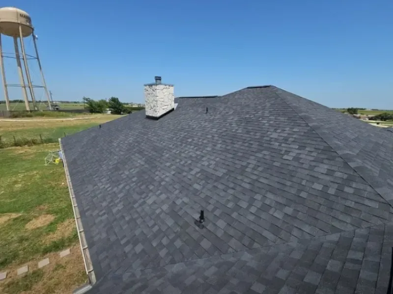 Dark gray asphalt shingle roof on a house, with a chimney. A water tower is in the background on a sunny day.