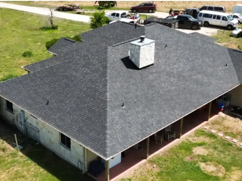 Dark gray asphalt shingle roof on a single-story house with chimney and porch; vehicles parked in yard.