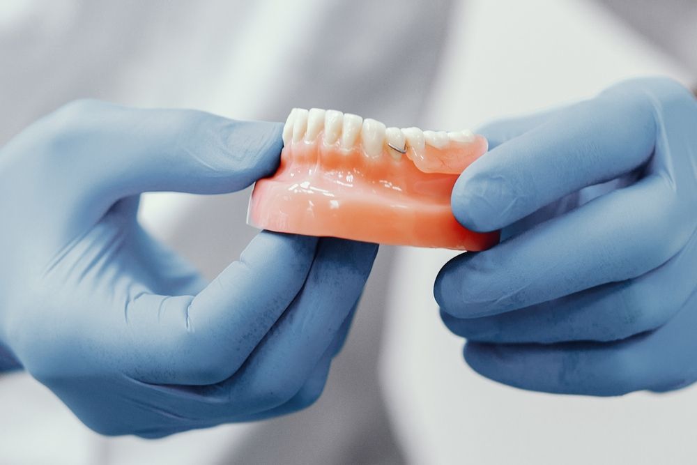 A Person Wearing Blue Gloves is Holding a Denture in Their Hands — Dental Prosthetics By Richard & Cindy In Oxenford, QLD