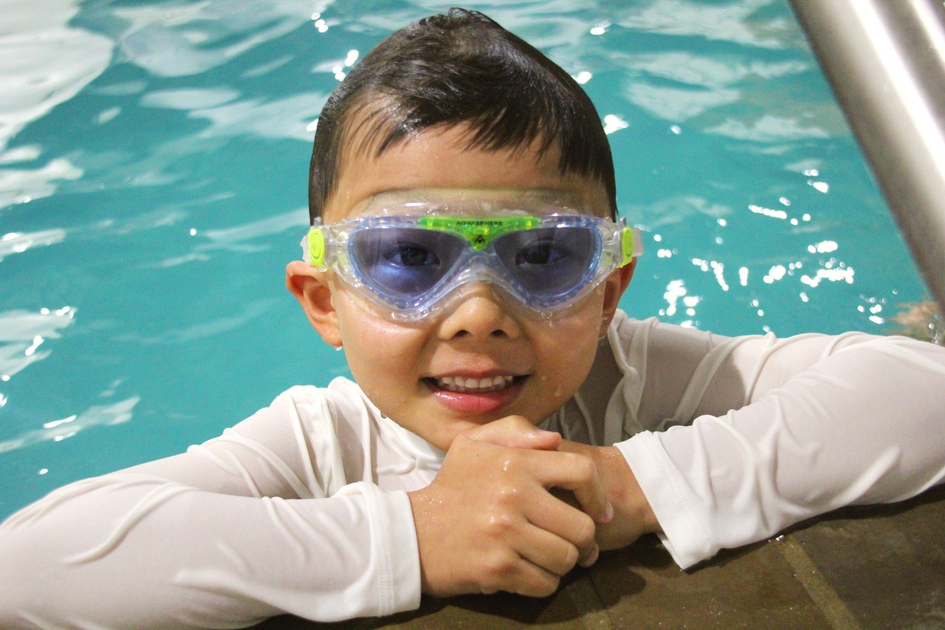 A young boy wearing goggles is leaning on the edge of a swimming pool.