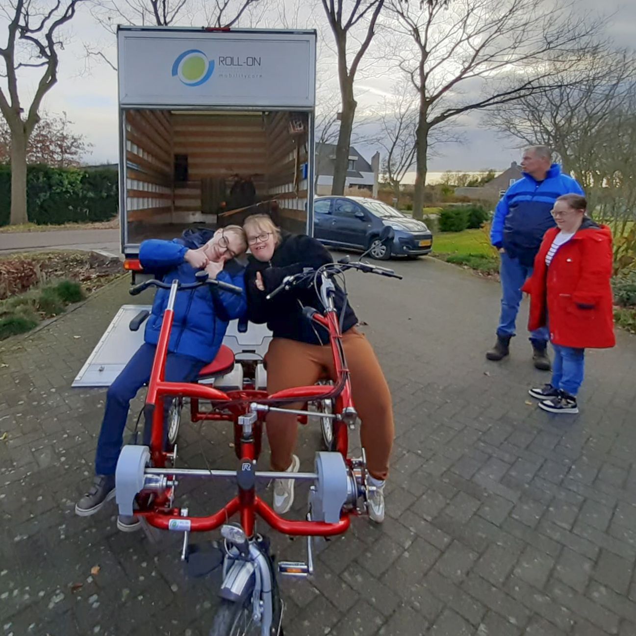 Two people on a red recumbent bike pose near a moving truck; two others watch nearby.