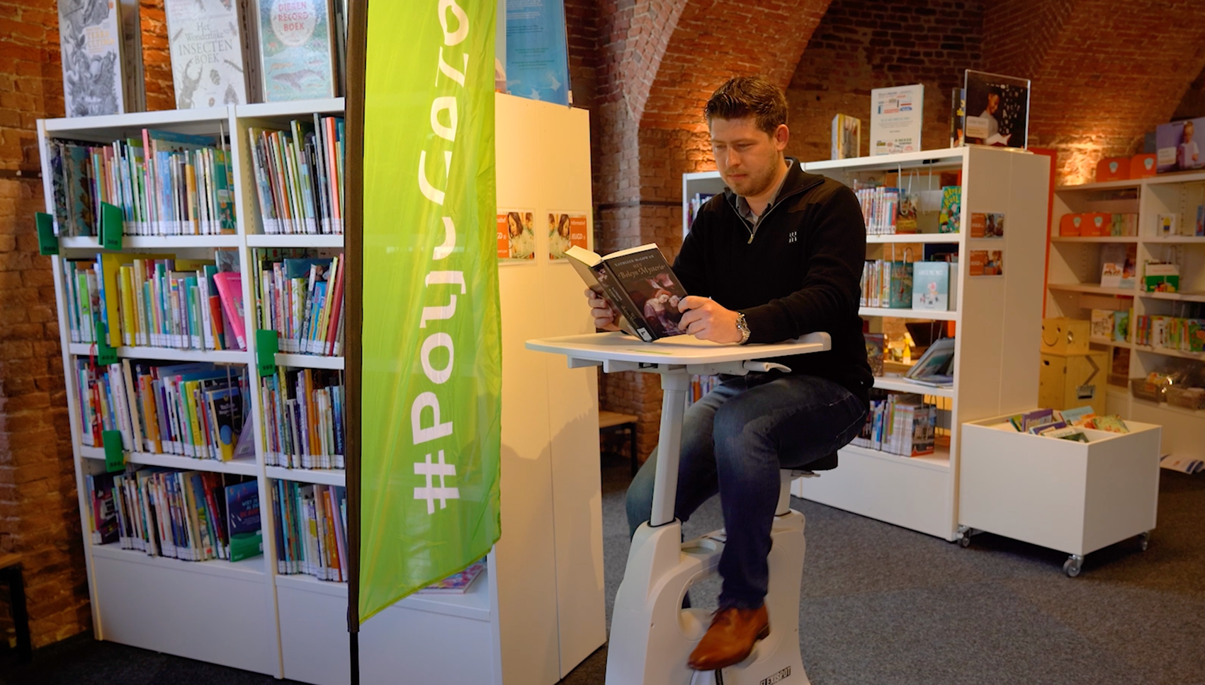 Een man zit op een leesfiets een boek te lezen in de bibliotheek van Oudenbosch.