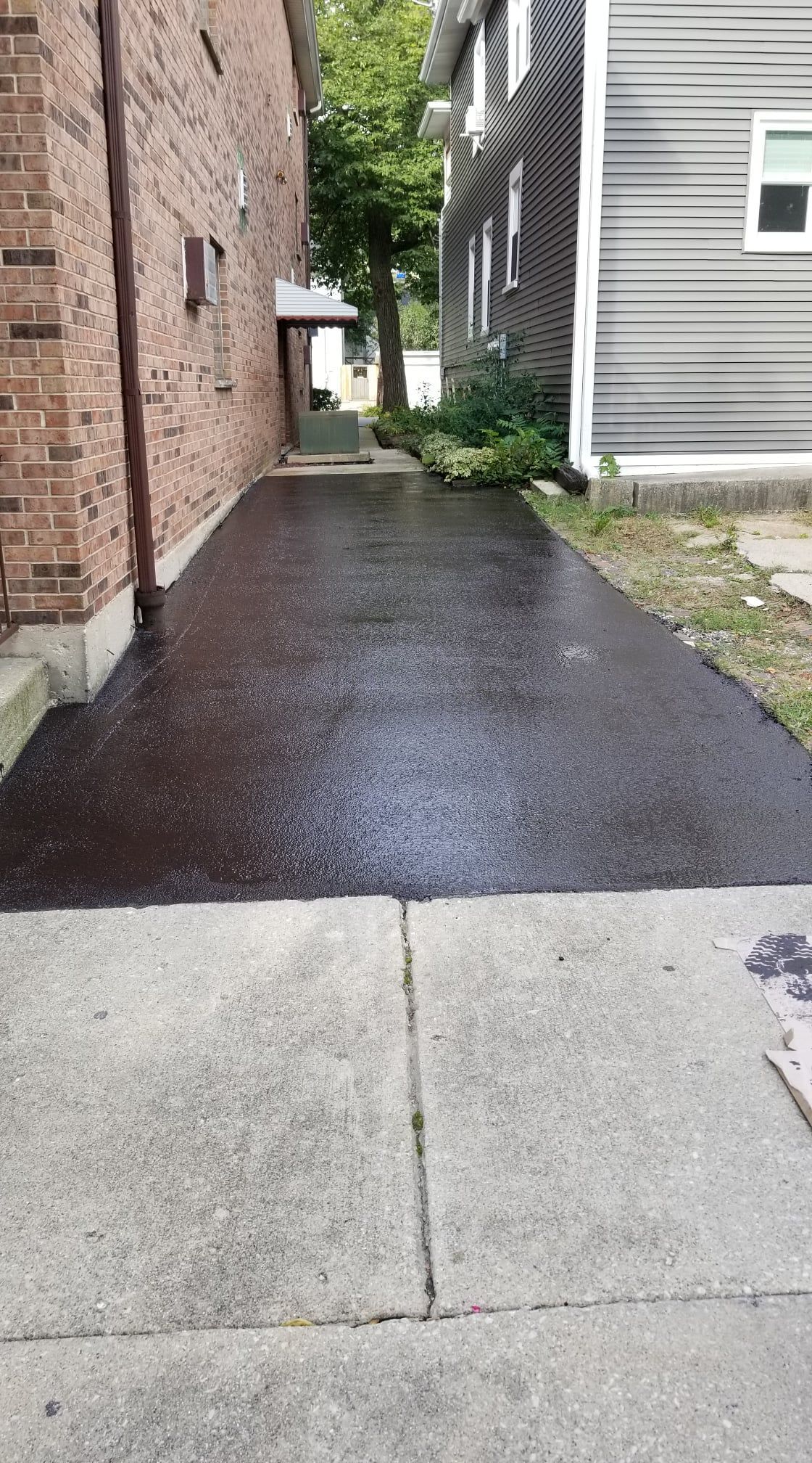 Asphalt driveway between brick building and gray-sided house. Wet surface, sidewalk in foreground.