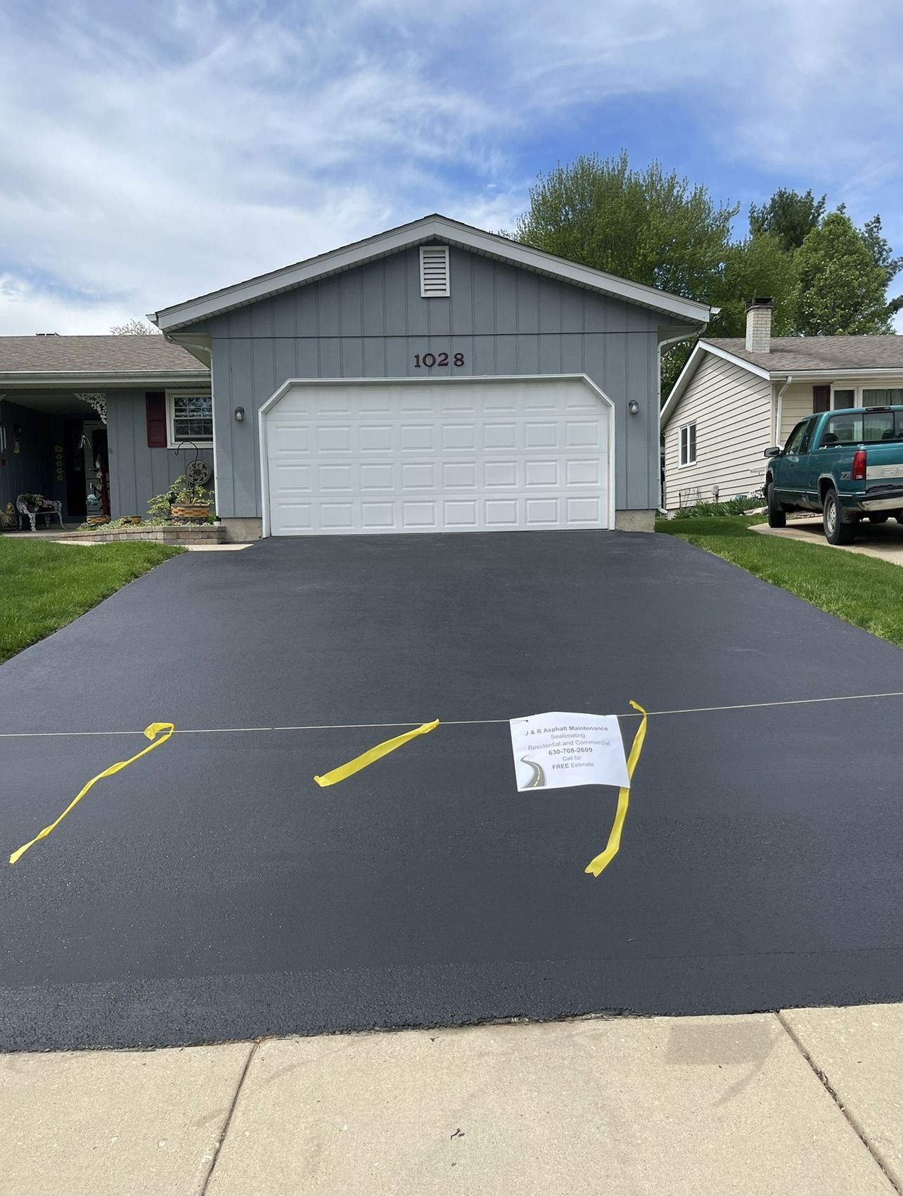 Newly paved black asphalt driveway in front of a blue-gray house with a white garage door, yellow markings, and a white sign.