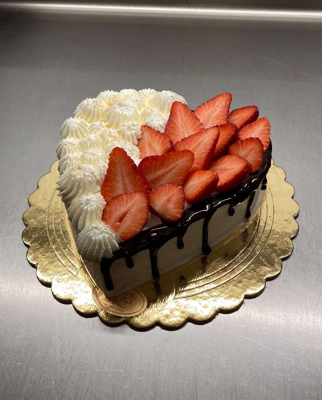 Torta a forma di cuore con panna montata, fragole e gocce di cioccolato su un centrino dorato.