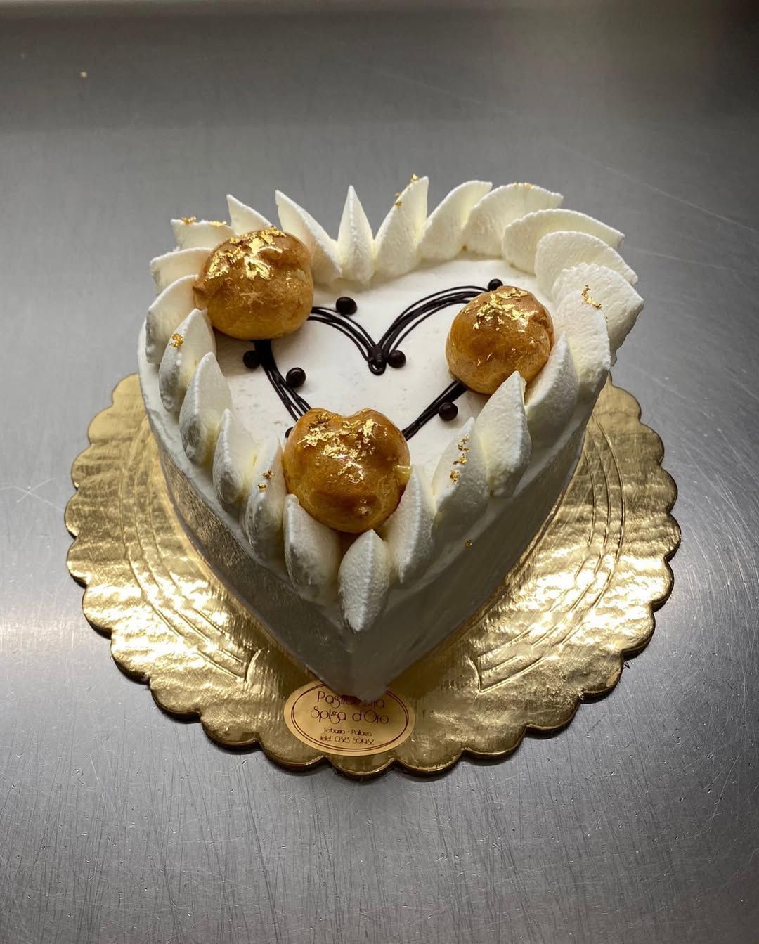 Torta a forma di cuore con glassa bianca, dettagli dorati e bignè, su un piatto dorato smerlato.