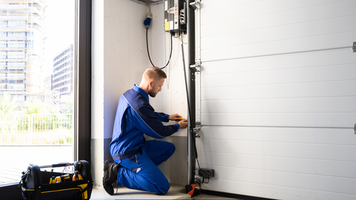 A man is kneeling down in a garage working on a garage door.