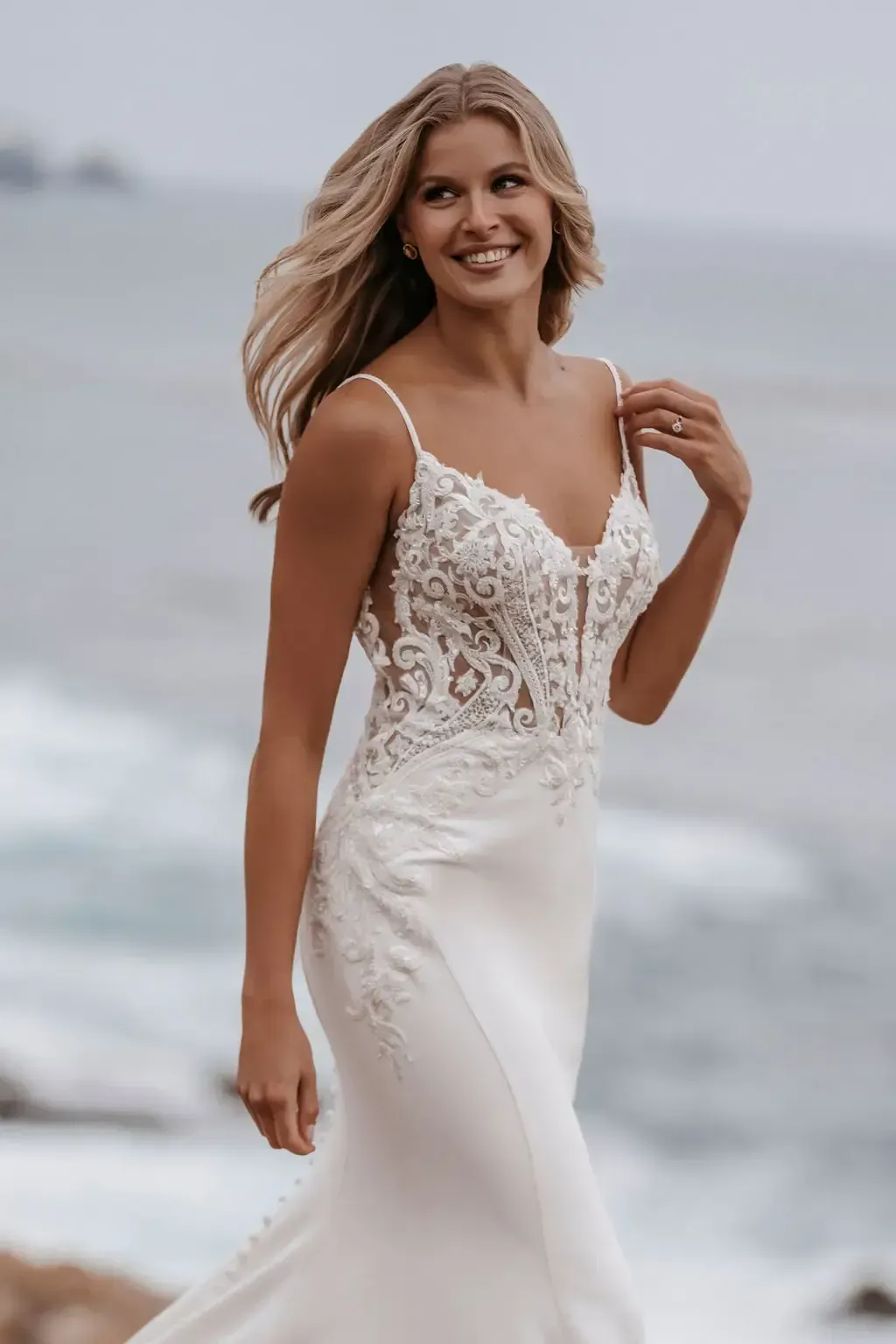 Woman in a white wedding dress smiles, standing near the ocean.