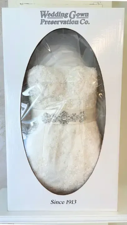 Wedding dress preserved in a box, visible through an oval window. 