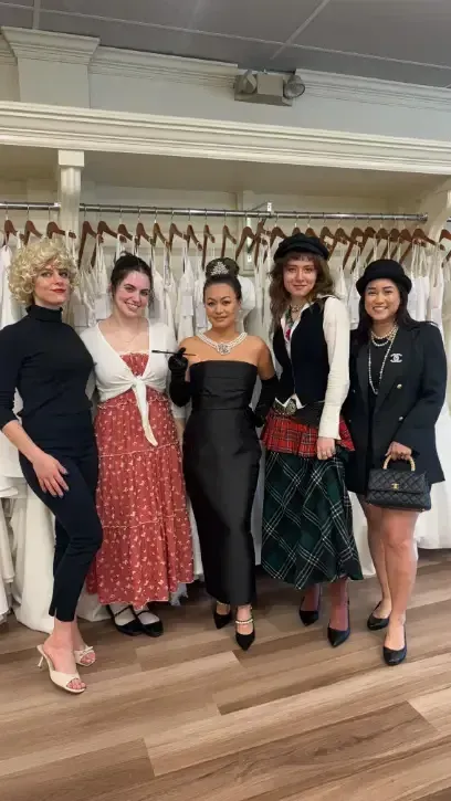 Five people in various outfits pose in a bridal shop. Dresses hang in the background.