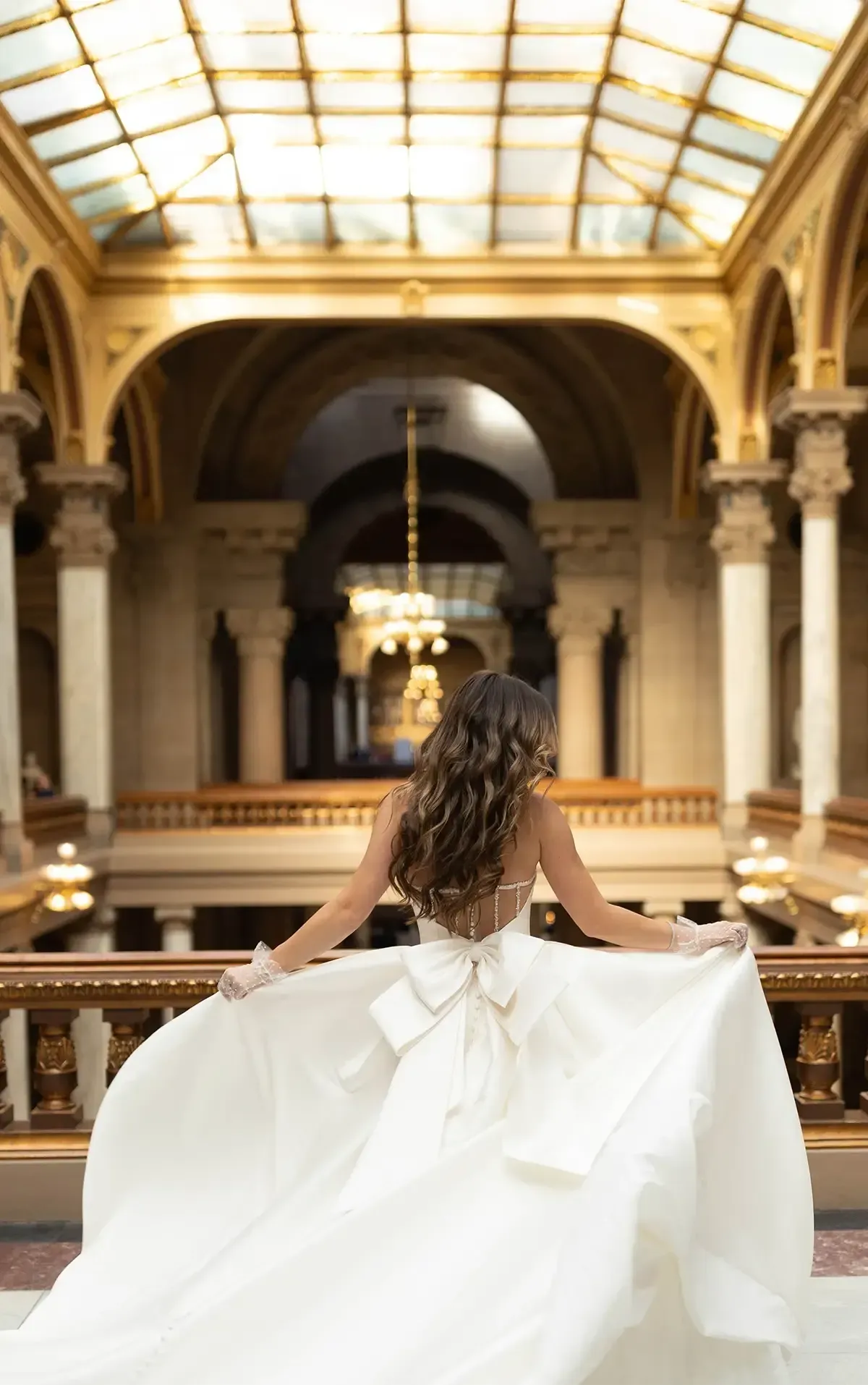 Woman in a white gown stands on a balcony, arms outstretched, looking towards a grand hall with marble columns and glass ceiling.