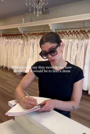 Woman signing a notebook at a boutique counter, with dresses hanging behind her.