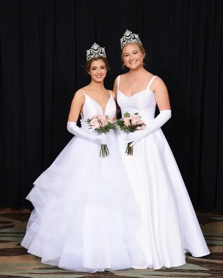 Two women in white ball gowns, tiaras, and gloves hold bouquets. They stand in front of a black backdrop.