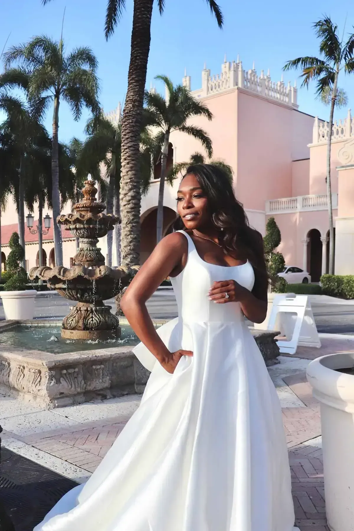 Woman in a white dress stands near a fountain, with a pink building and palm trees in the background.