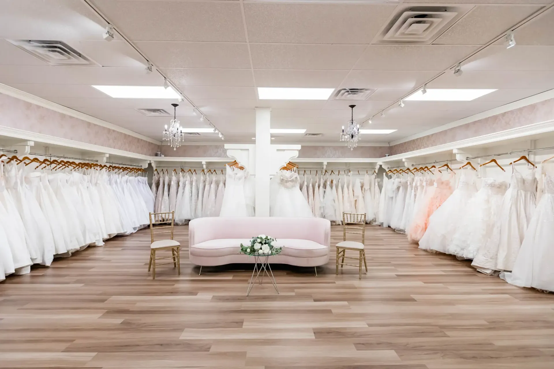 Bridal shop interior with many white wedding dresses on display, a pink sofa, and wooden floor.