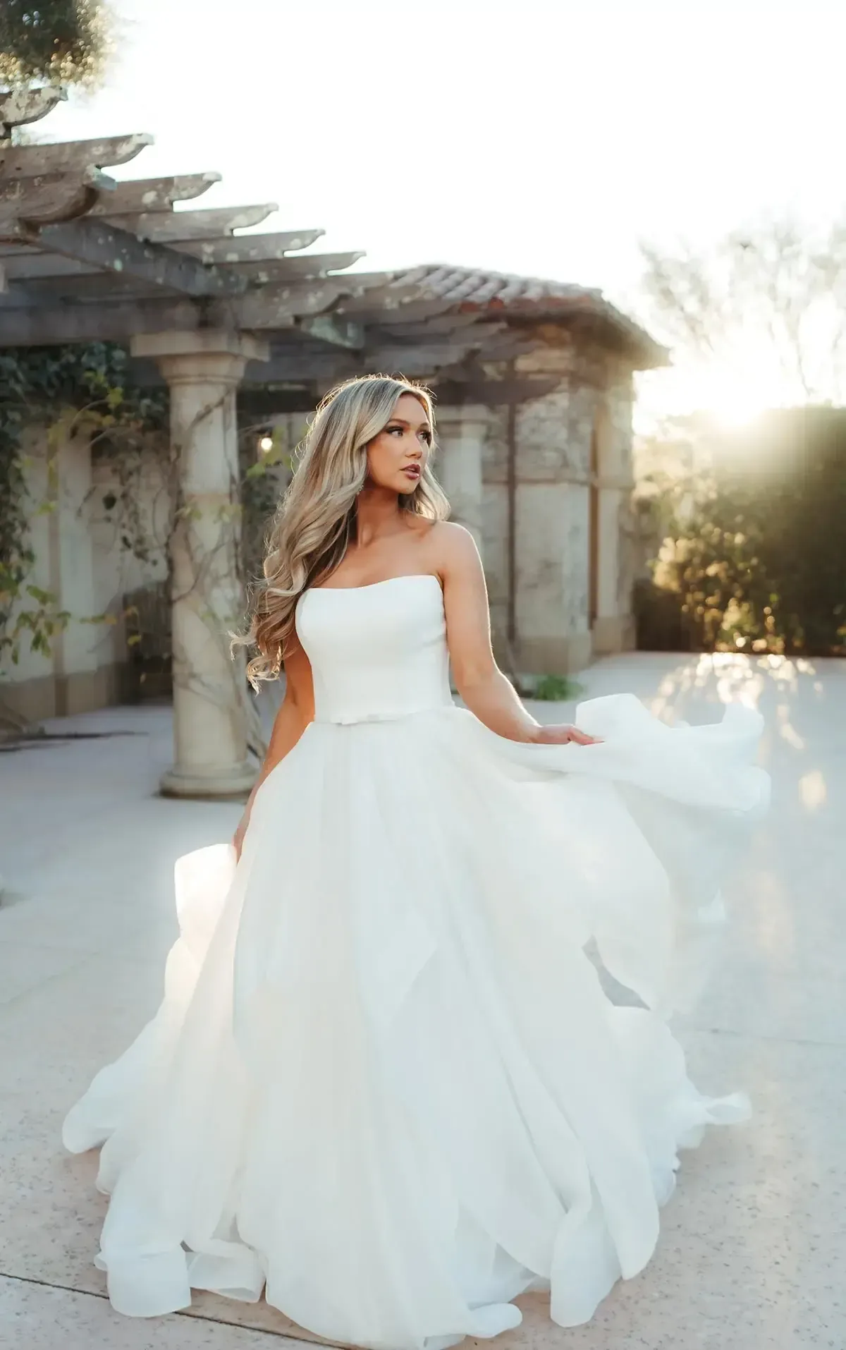 Woman in white strapless gown poses outdoors. Flowing skirt, long blonde hair, sunlit garden backdrop.