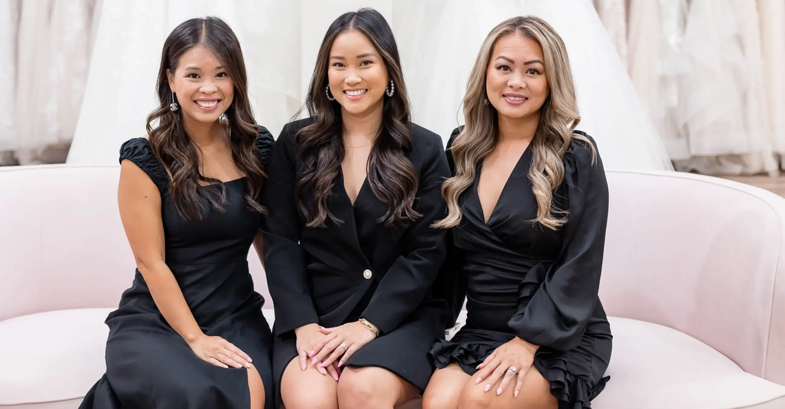 Three women smiling on a pink couch in a bridal shop, dresses in the background.