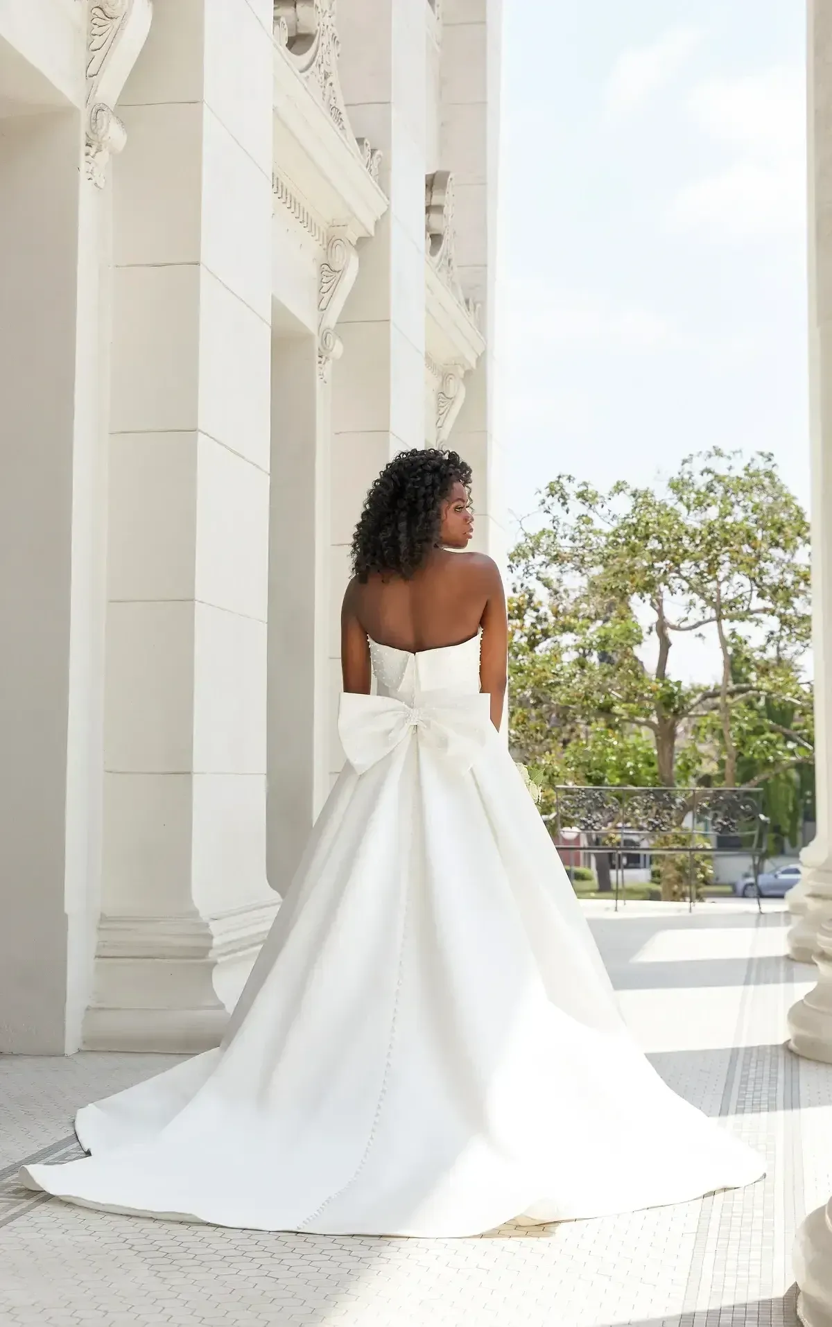 Woman in a strapless white wedding dress with bow, standing outside under tall columns.