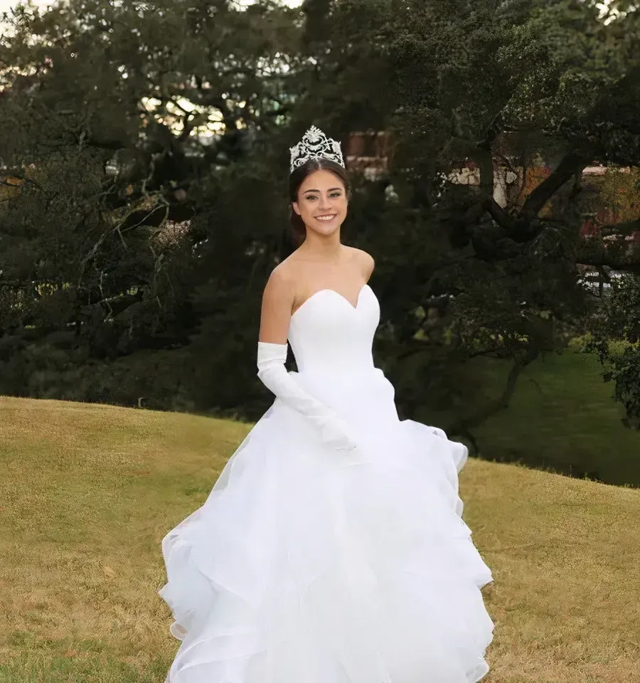 Woman in a white ball gown and tiara smiles outdoors, in front of trees.