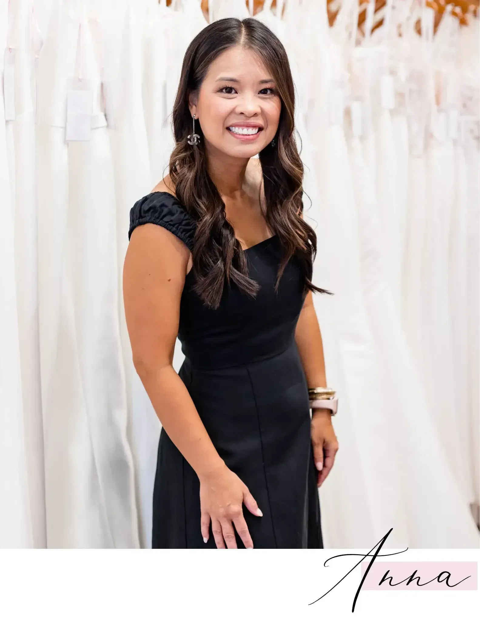 Woman in a black dress smiling in a bridal shop, rows of white dresses in the background.