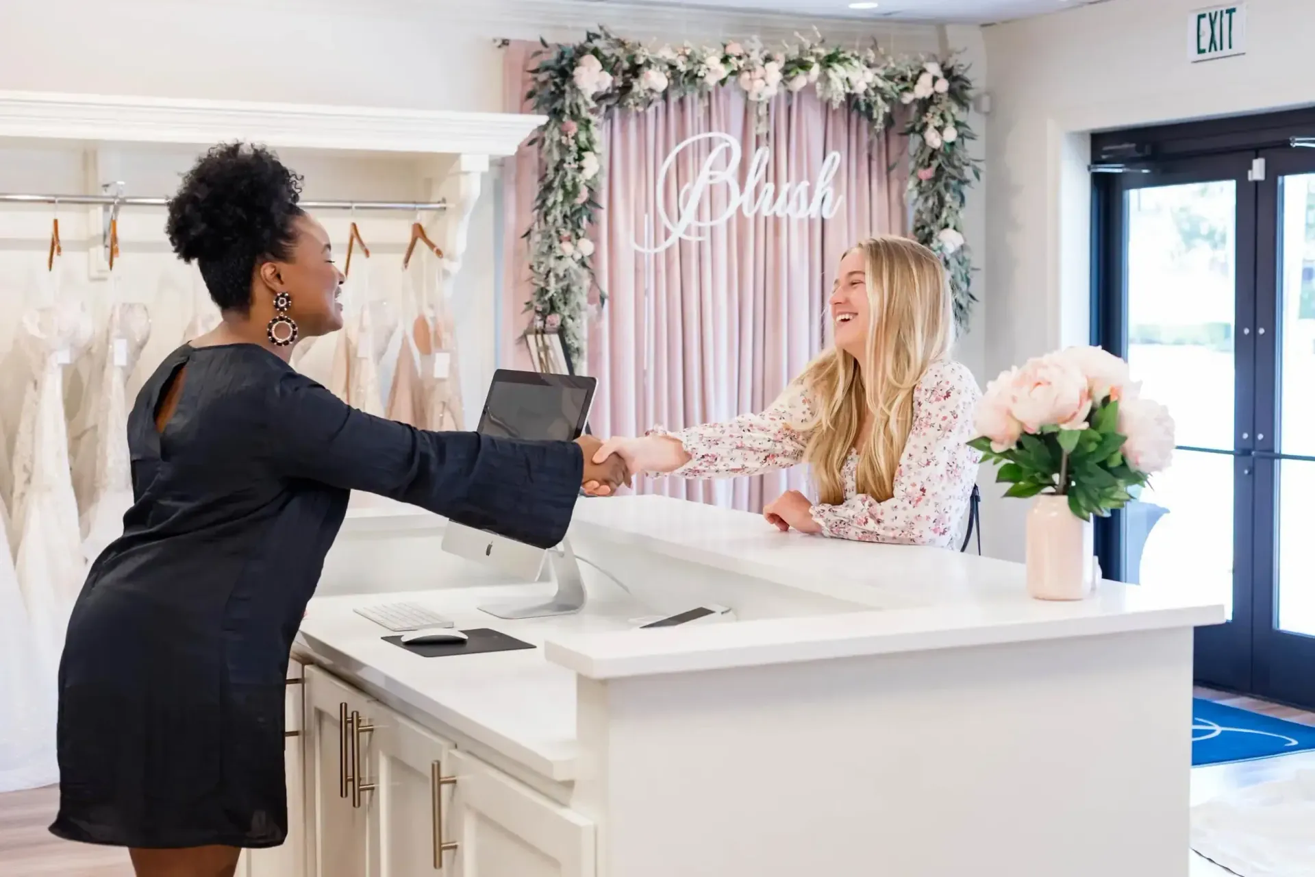 Woman in black dress shaking hands with woman at a white reception desk, smiling in a bridal shop.