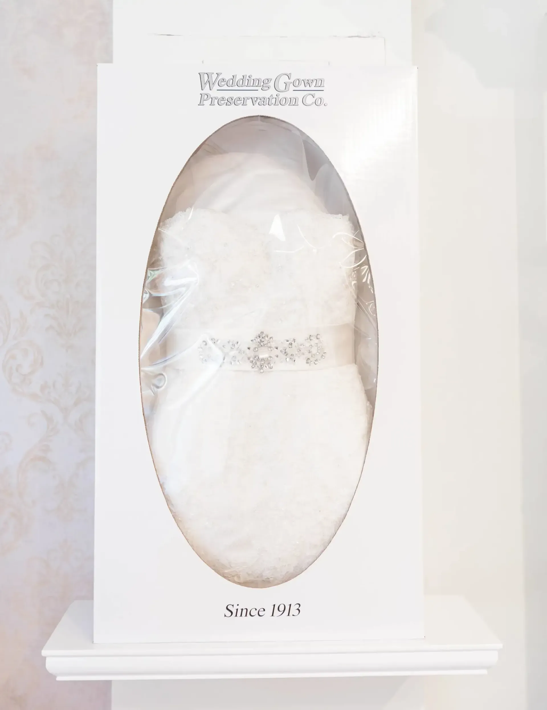 Wedding dress in white box with clear window on a shelf.