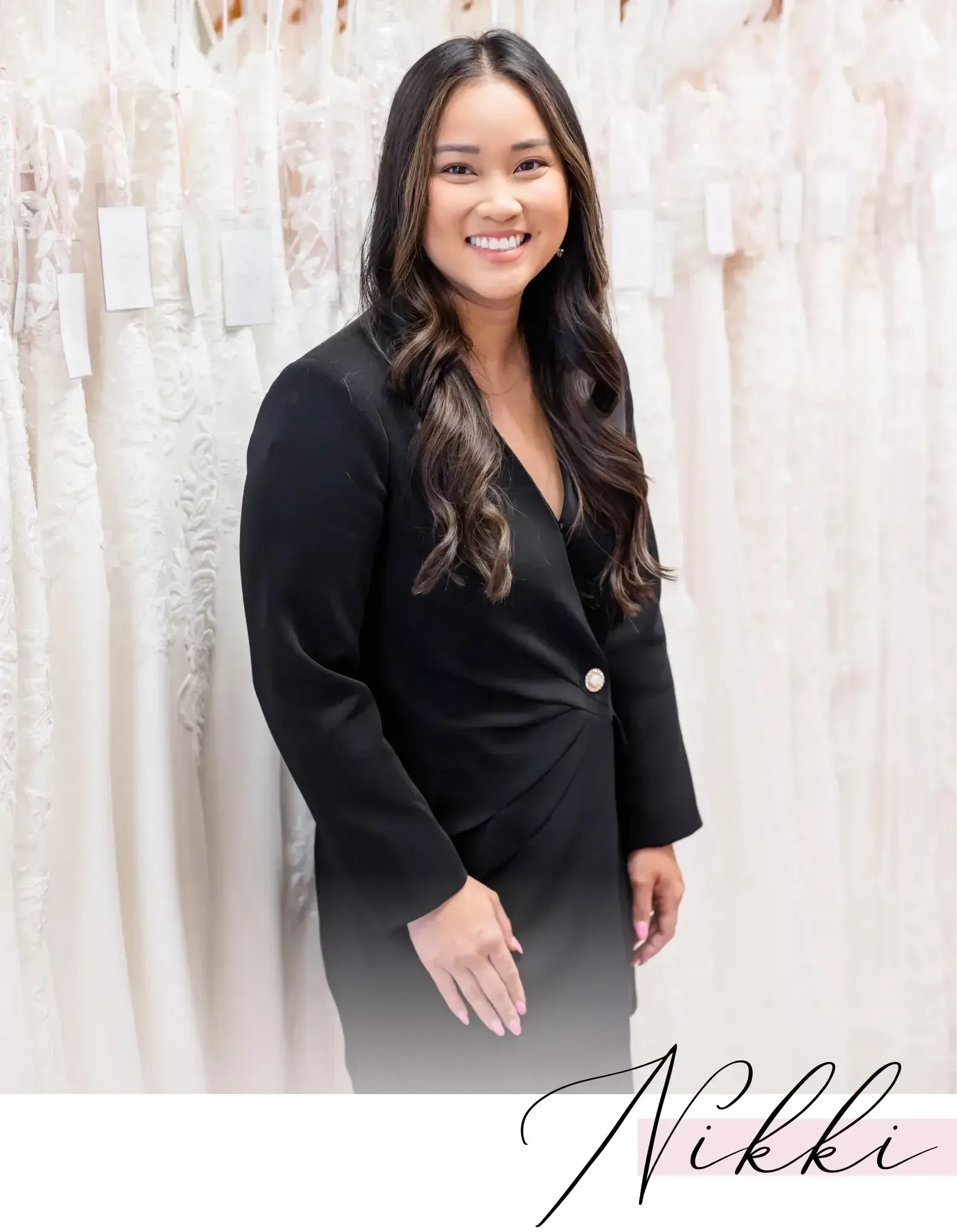 Woman smiling in a black jacket, posing in front of wedding dresses.