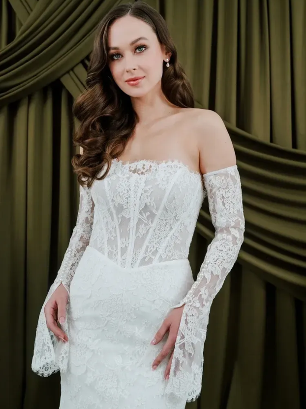 Woman in a white lace off-the-shoulder wedding dress with long sleeves, posing in front of olive green drapery.