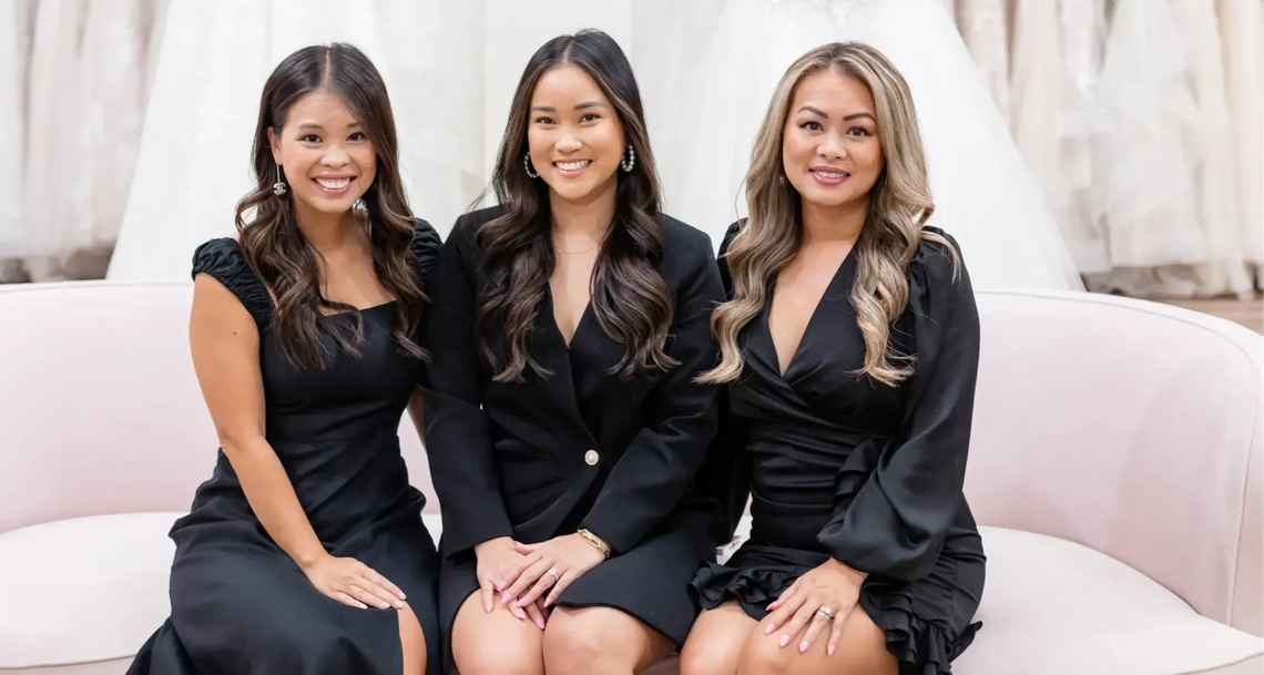 Three women smiling on a pink couch in a bridal shop, dresses in the background.