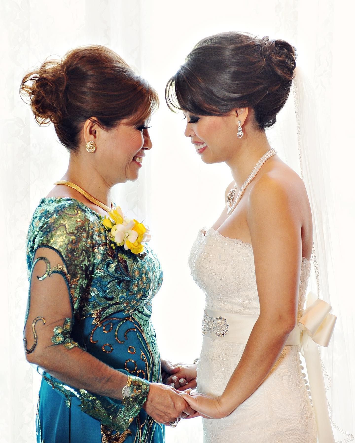 Bride and mother touch foreheads, smiling. Bride in white dress, mother in teal gown, by a window.