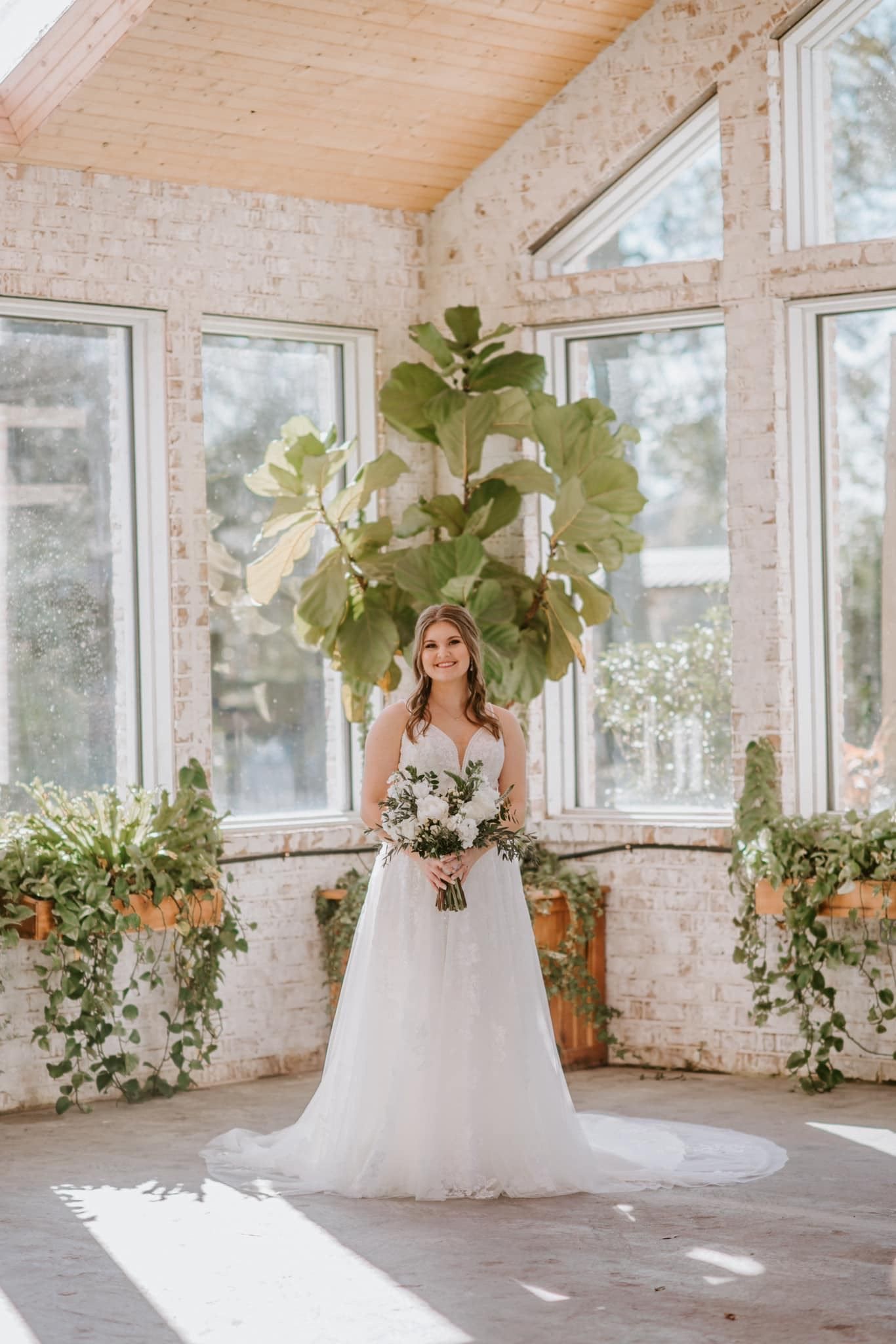 Bride in white dress, holding bouquet, in a bright sunroom with large plant.
